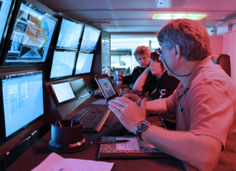 Team members on watch stand in the R/V Falkor’s science control lab, where the monitors display real-time multibeam, magnetometer, and gravimeter data streams. Watch standers are responsible for monitoring and logging the quality of the incoming data, communicating with the bridge if course and speed adjustments are warranted, transmitting data to the processing computers in the adjoining room, and conducting sound velocity profiles. Credit: Daniel Wagner