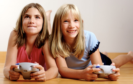 Two girls lie on their stomachs on the floor, holding video game controllers and looking up.