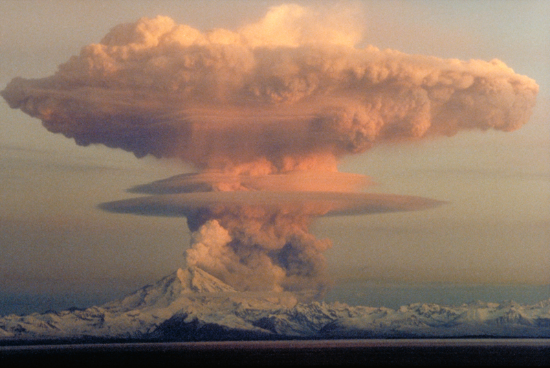A plume of ash ascends in a massive column above a snow-capped volcano.