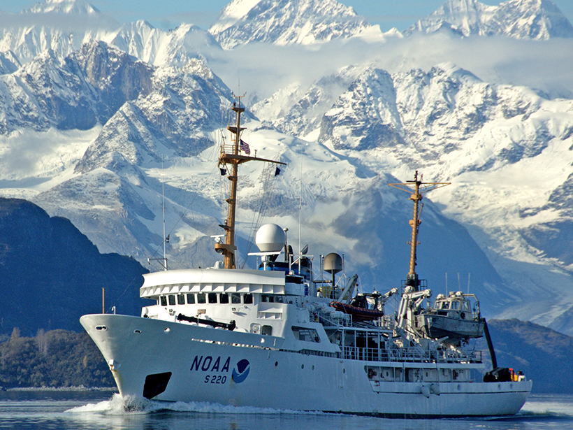 A NOAA ship sails through a bay beneath snow-covered mountains.