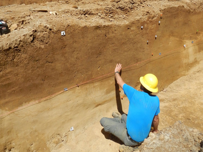 Seated man in a hard hat assesses sediment patterns in a dug trench