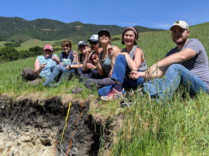 College students sit on the rim of a soil pit on a grassy hill.