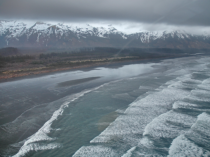 Aerial photo of a dark sand beach at low tide with snow-capped mountains in the background
