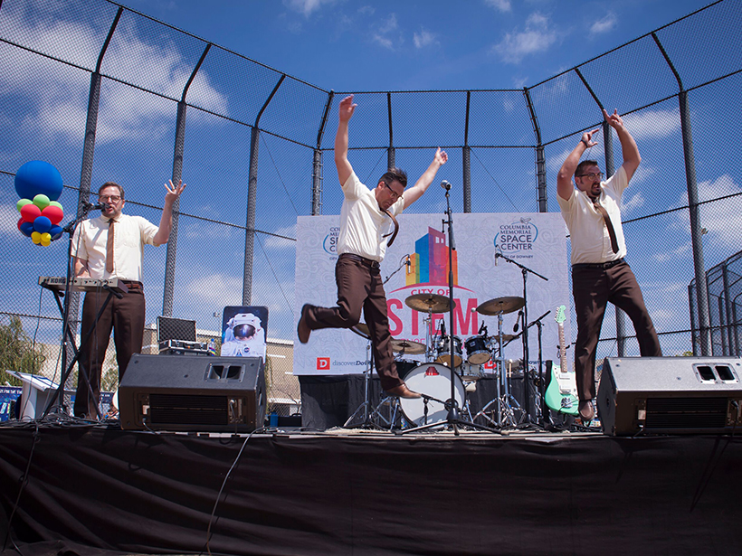 Three white guys in shirtsleeves perform on an outdoor stage.