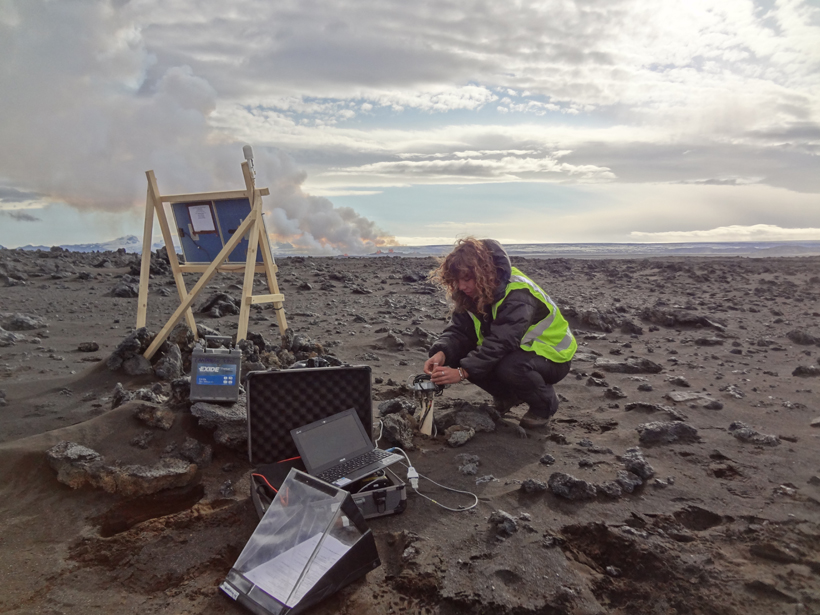 A woman conducts experiments with a laptop in a lava field while volcanoes erupt in the distance.