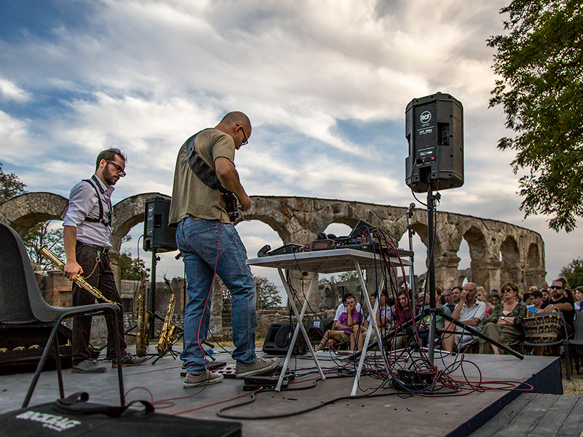 Two musicians play on stage in front of an outdoor audience at an ancient Roman theater.