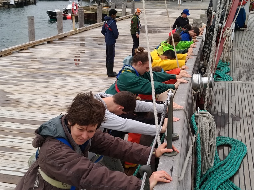 Female students pushing a wooden sailboat away from a dock in Woods Hole, Mass.