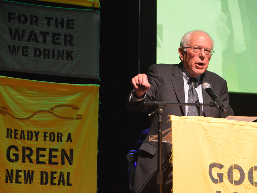 Photo of Bernie Sanders speaking at a podium with Green New Deal banners