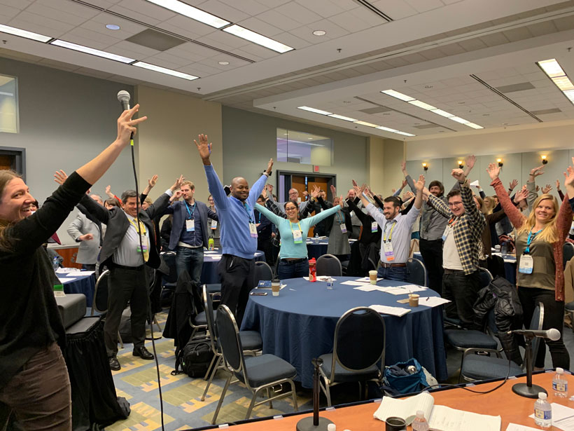 A room full of cheerful people stand during a science communication workshop.