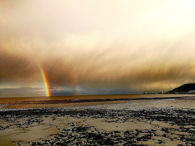 Clouds and rainbows hang over a broad beach on Britain’s west coast.