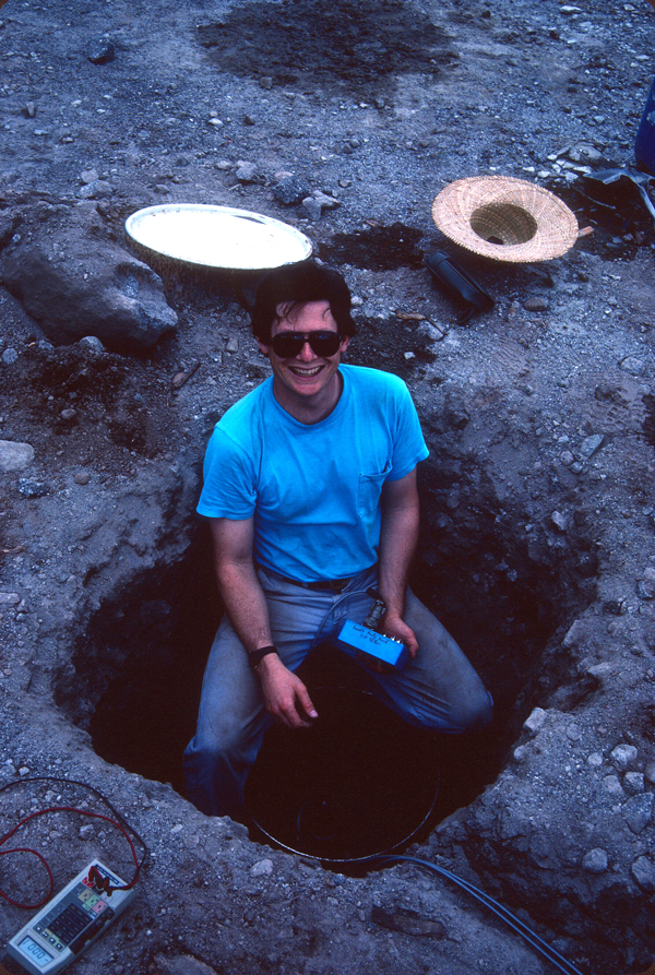 A smiling volcanologist installs a tiltmeter in a hole in the ground.