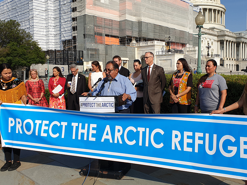 A small group of people speak at a podium in front of the Capitol with the banner “Protect the Arctic Refuge.”