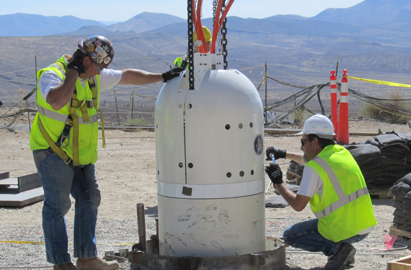 Two men in hardhats and safety gear lower a large canister into a borehole. Two men in hardhats and safety gear lower a large canister into a borehole.