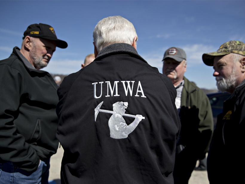 A man in a United Mine Workers of America jacket talks, back to the camera, with other men.