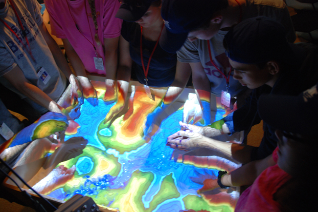 Student hands create geological features in an augmented reality sandbox. Student hands create geological features in an augmented reality sandbox.