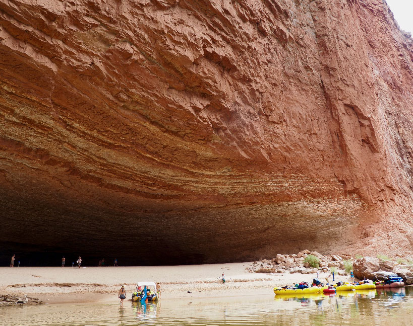 A half dozen rafts are tied up in front of a huge cavern scooped out of Redwall Limestone in a bend in the Colorado River.