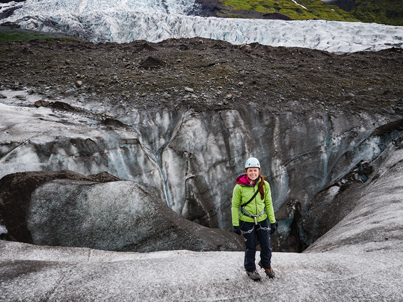 A woman outfitted in climbing gear stands on a dirty glacier.