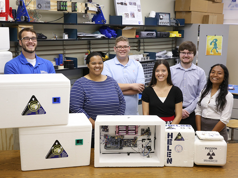 College students smile with open and closed boxes with tech equipment, labeled HELEN