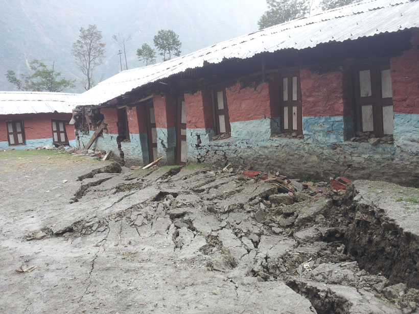 Large earthen rupture foregrounds a brightly colored school building damaged by an earthquake.