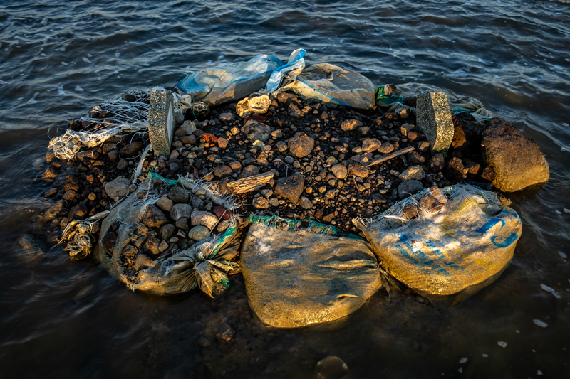 Frayed plastic bags of rubble anchor a drowned gravesite.