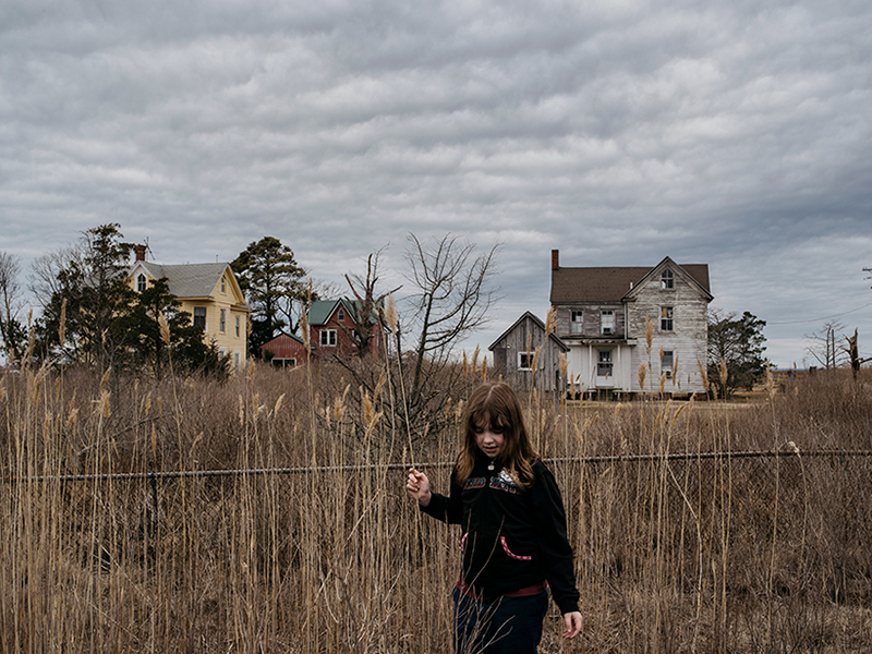 A second-grade girl beside a chain-link fence and tall grass.