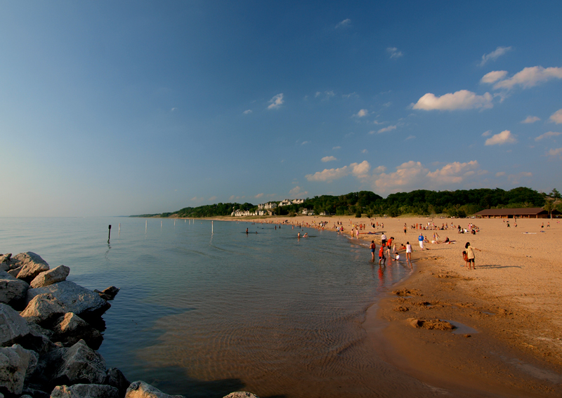 Dozens of beachgoers enjoy a sandy beach and calm swimming area on Lake Michigan.