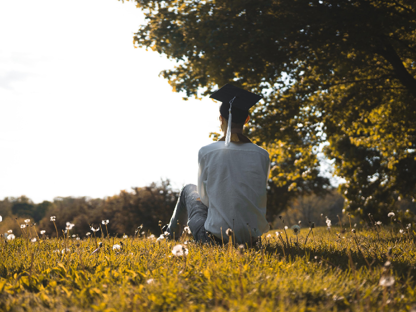 Person in a graduation cap sits with their back to the camera amid a field of dandelions.