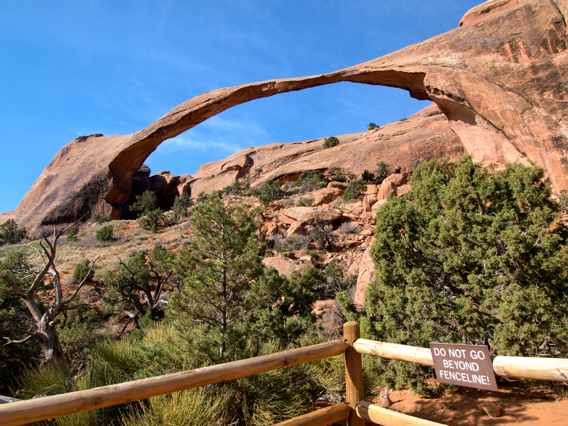 Landscape Arch arcs across a fence and sign warning “Do not go beyond fenceline!”