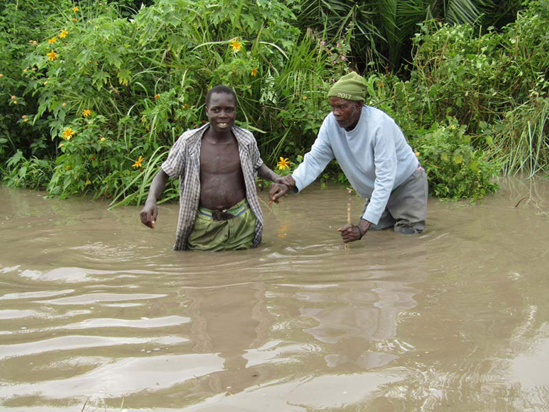 Two men stand in floodwaters above their knees, the younger man holding the arm of the older man, who is leaning on a cane in the water. Two men stand in floodwaters above their knees, the younger man holding the arm of the older man, who is leaning on a cane in the water.