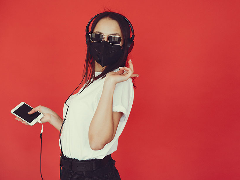 A woman in a respiratory mask and sunglasses listens to music on headphones against a bright red backdrop