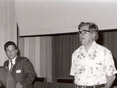 Black-and-white photo of Orson Anderson standing and speaking in front of a drop-down slide screen