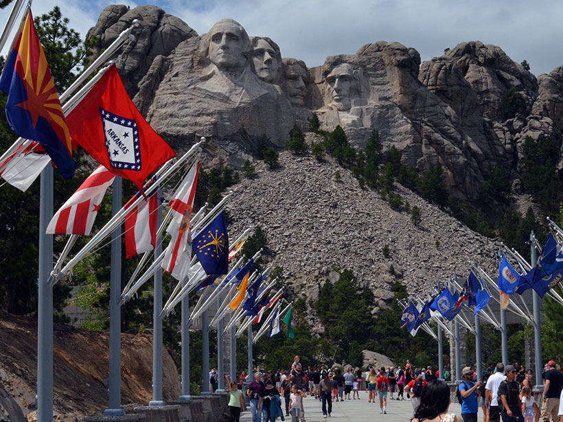 Four faces carved in granite stand above an apron of crushed rock overlooking state flags along the Avenue of Flags at Mount Rushmore National Memorial.