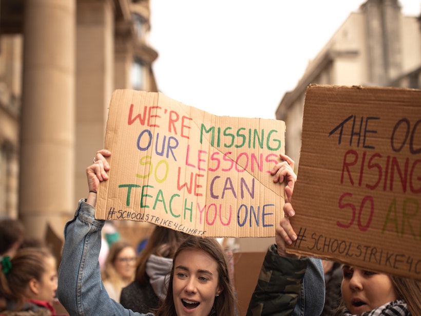 Young woman holds a sign saying “We’re missing our lessons so we can teach you one.”