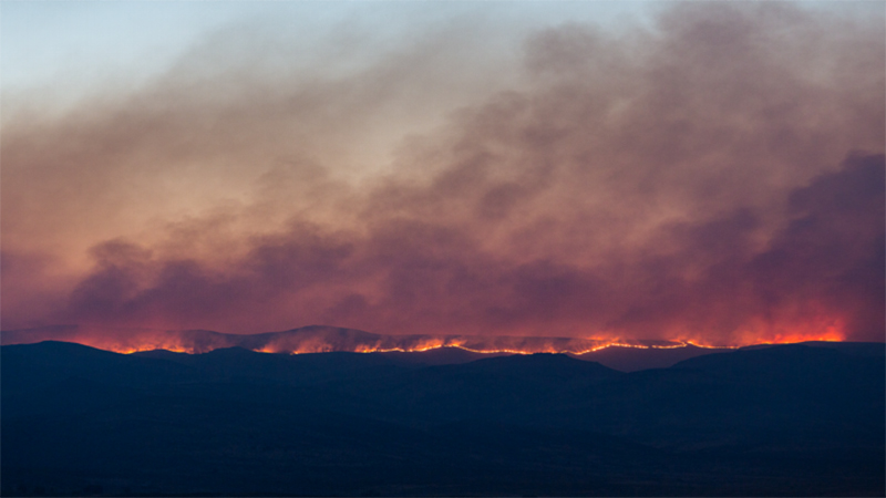 Smoke rises from a line of wildfire burning the landscape in the distance.