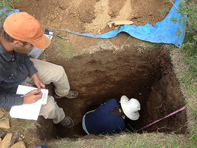 A researcher crouches in a deep pit collecting soil samples while another takes notes.