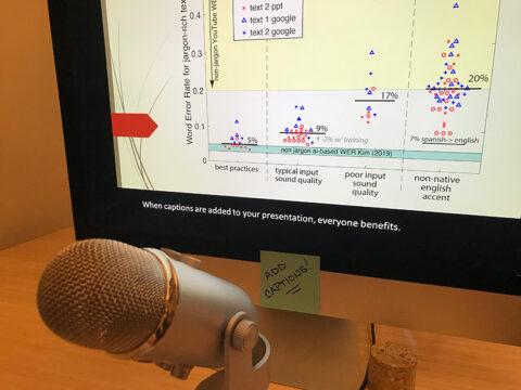 A large microphone sits in front of a computer monitor displaying a graph on captioning