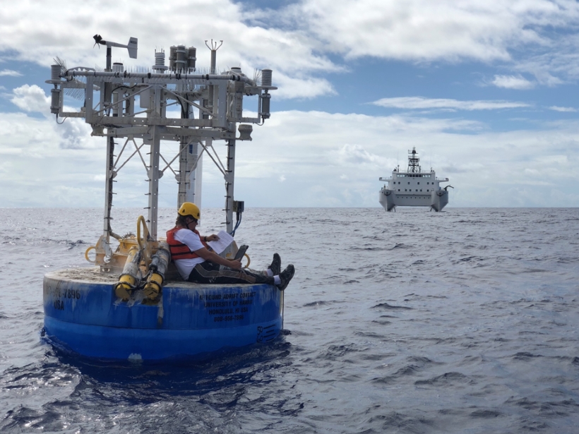 A researcher checks a carbon sensor manual on a floating research buoy while a research ship waits in the distance