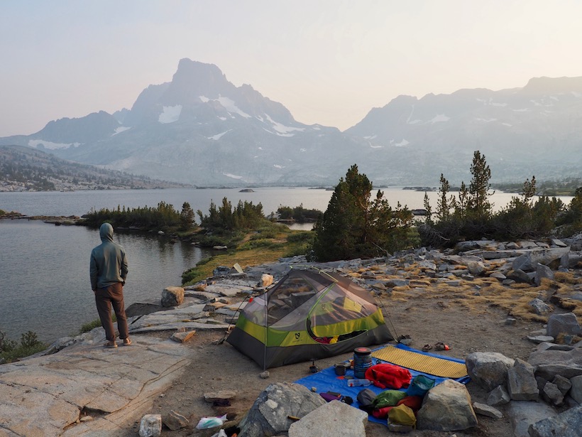 A hiker stands next to a lakeside campsite, looking across Thousand Island Lake to Banner Peak, which is cloaked in smoke.