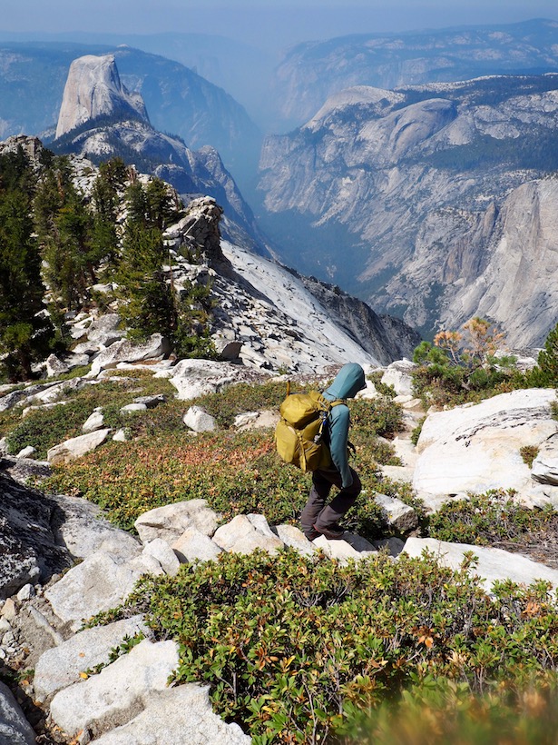 A backpacker makes their way down Clouds Rest on a stone staircase, as Half Dome towers over Yosemite Valley.