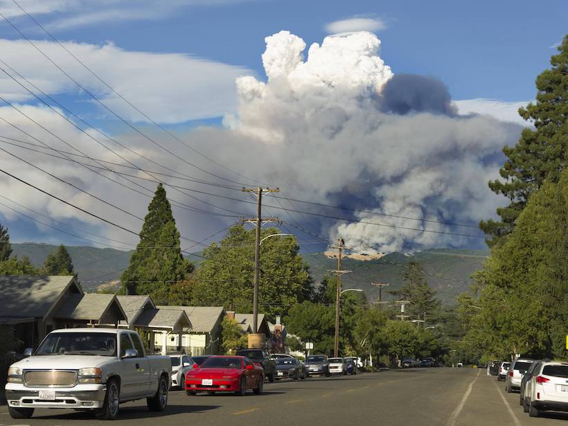 Smoke billows in the distance from a mountain near Ukiah, Calif., as motorists drive down a street in the foreground.