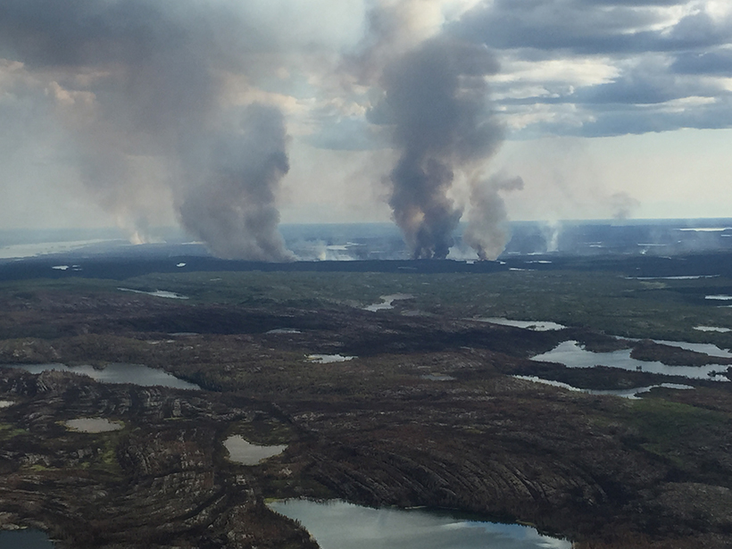 Smoke rises from a singed landscape, meeting the clouds above a swath of boreal forest punctuated by lakes.