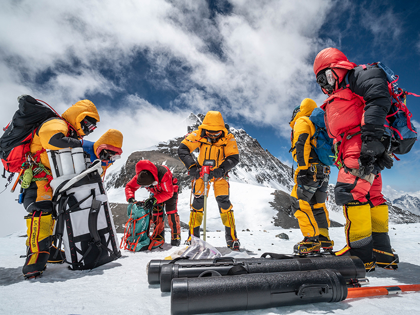 Six people in bright snowsuits and goggles drill an ice core on Mount Everest with mountains and clouds in the background.
