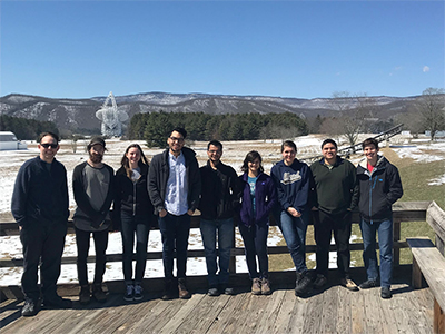Nine people stand in a line on a wooden deck with a radio telescope and snowy mountains in the background Nine people stand in a line on a wooden deck with a radio telescope and snowy mountains in the background