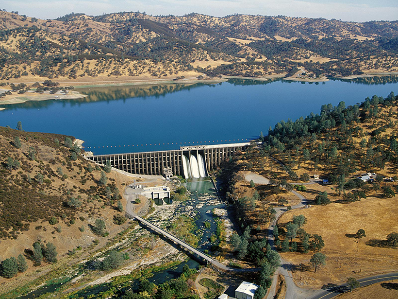 A blue reservoir lake surrounded by yellow hills with spotty dark green tree coverage flows into a hydroelectric dam.