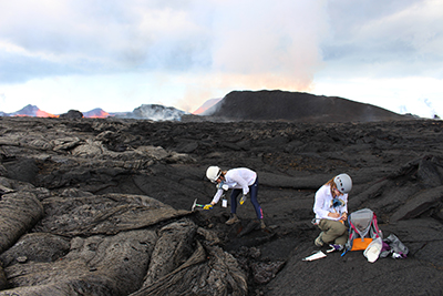 A researcher in a white shirt and hiking boots hammers at a field of freshly formed volcanic rock to collect a sample; another researcher takes notes nearby.