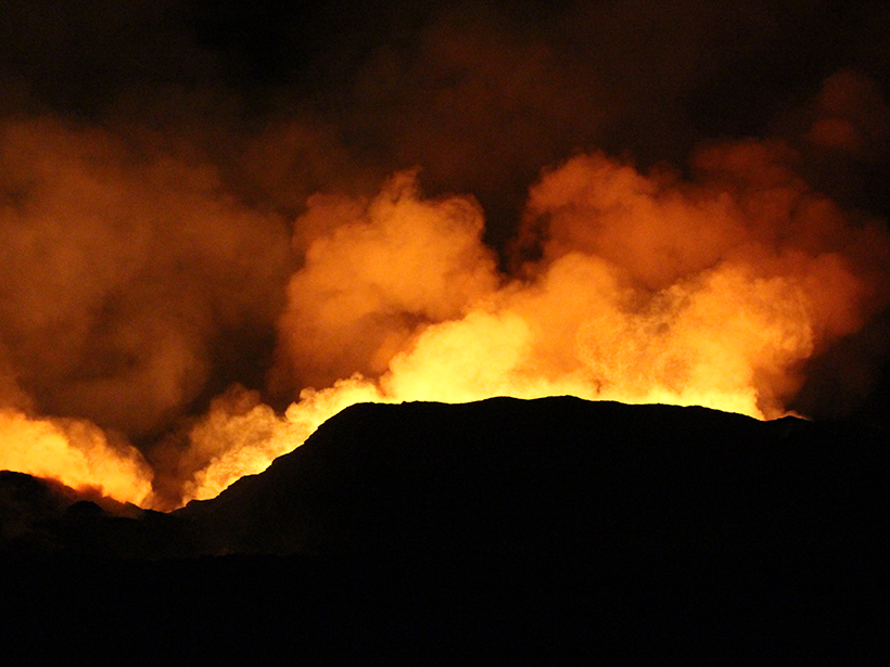 Image of orange and red smoke rising from behind the black banks of a lava channel at night