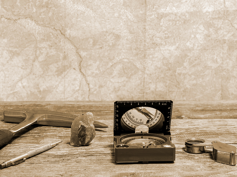 Rock pick, compass, and other tools of geology on a table with a map in the background