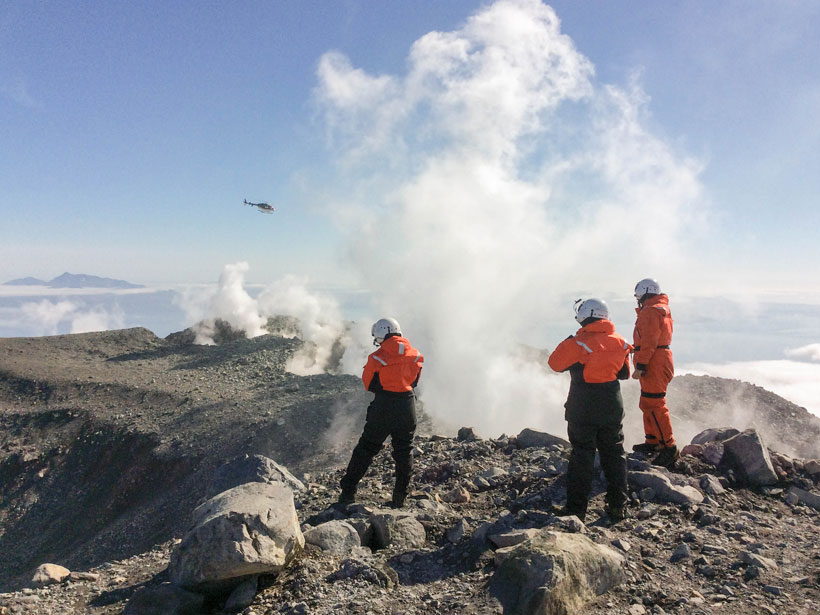 Scientists in flight suits stand atop the summit of a volcano with steam rising nearby and a helicopter in the distance