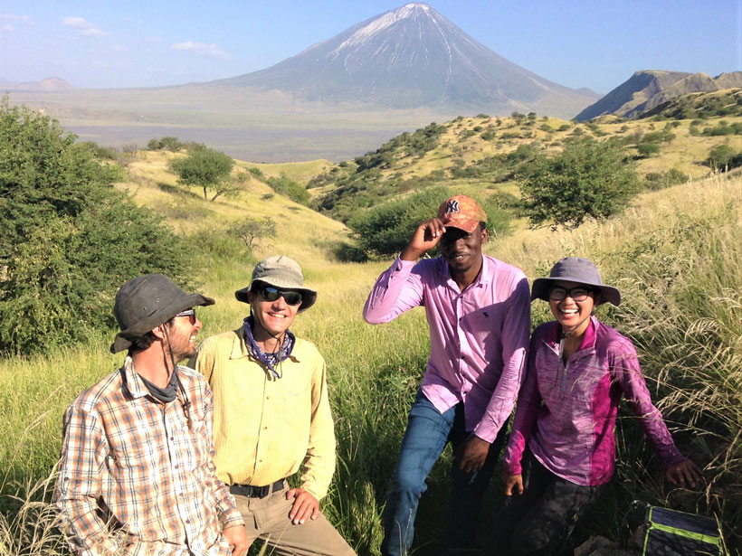 Researchers pose for a photo while standing in tall grass with a volcano rising in the background behind them