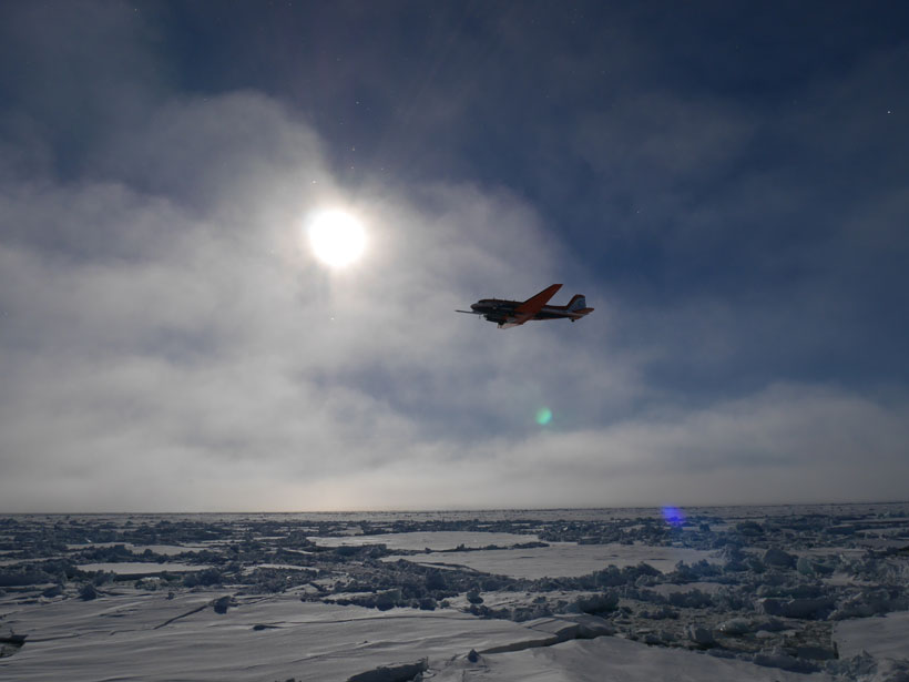 An airplane flies low over ice with the sun shining through clouds in the background